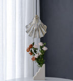 A Clérachic ribbed glass shell pendant, unlit and clear, hangs beside a white curtain with a vase of roses below it.