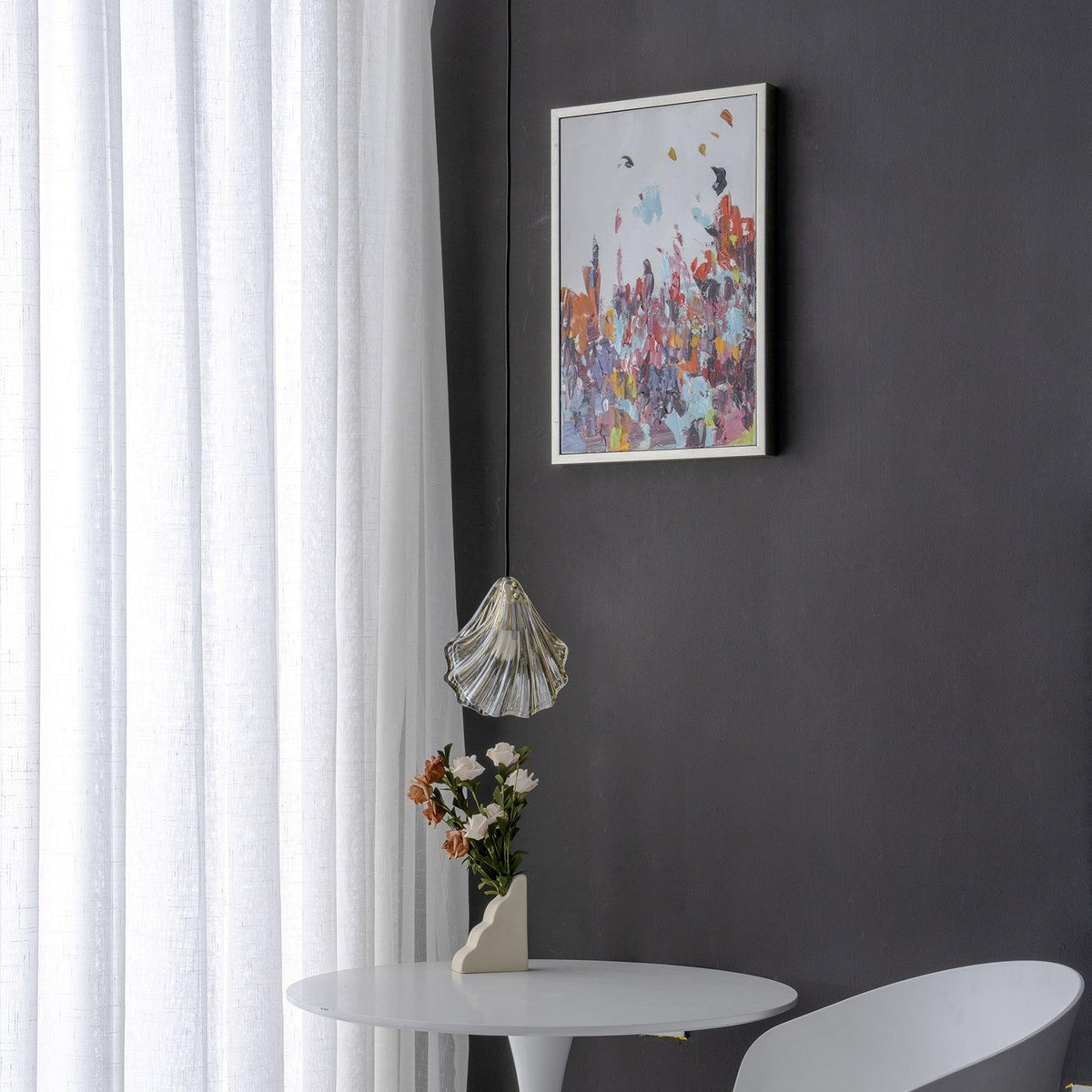 Wide view of a Clérachic shell pendant hanging low beside a framed abstract print, with a vase of flowers on a white table.