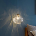 A lit Clérachic shell pendant with clear ribbed glass spreads fine rays of light across a blue wall above a grey pillow.