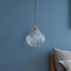 The same Clérachic shell pendant switched off, showing clear glass ribs and a brass fitting against a blue wall.
