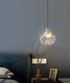 A Clérachic clear glass shell pendant hangs above a bedside shelf with a dried stem vase, against a grey-blue wall.