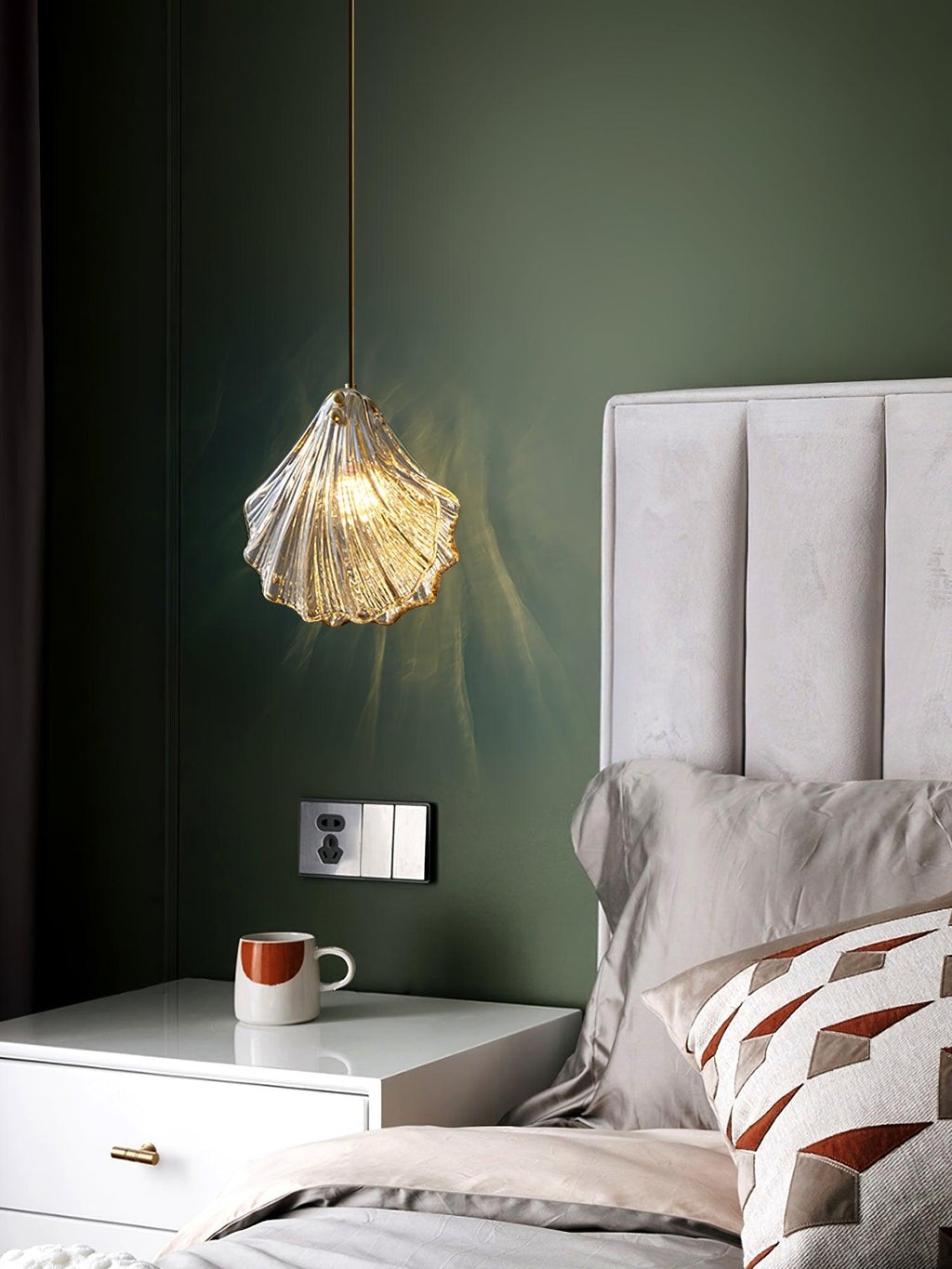 Against an olive-green wall, a Clérachic ribbed glass pendant hangs above a white bedside table with a mug and a cushioned bed.