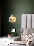Against an olive-green wall, a Clérachic ribbed glass pendant hangs above a white bedside table with a mug and a cushioned bed.
