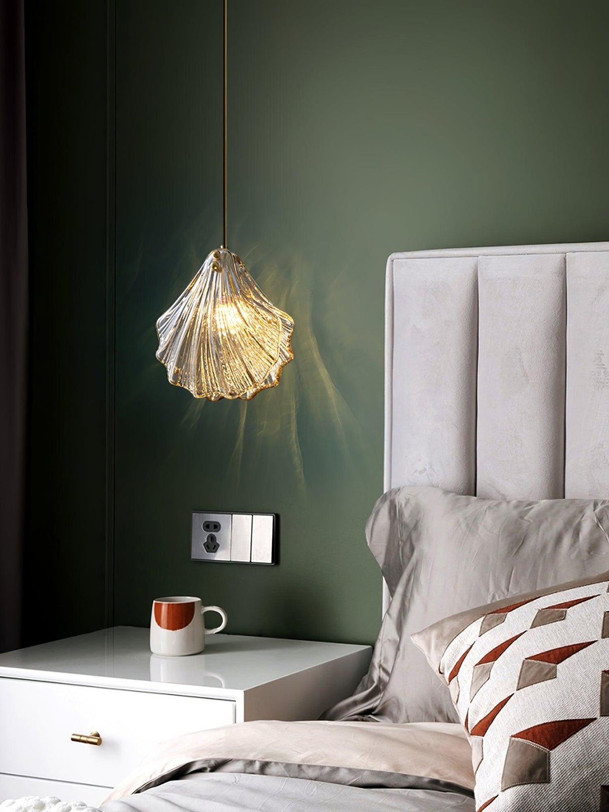 Against an olive-green wall, a Clérachic ribbed glass pendant hangs above a white bedside table with a mug and a cushioned bed.