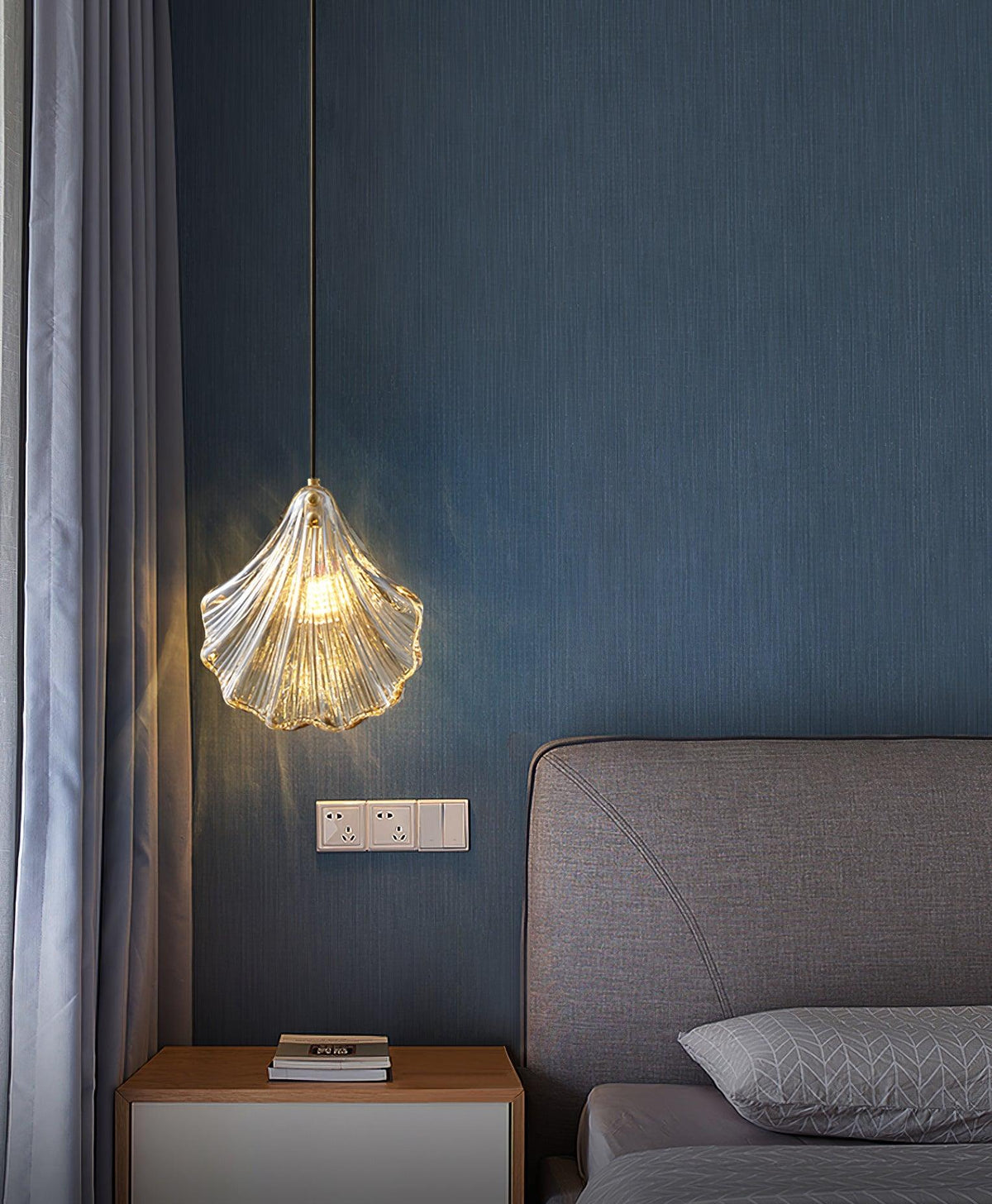 A Clérachic shell pendant hangs by a grey curtain and a grey upholstered headboard, against a deep blue textured wall.