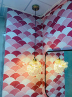 Two Clérachic shell pendants with golden glass hang in a corner tiled in pink and white scallops, one reflected in a mirror.