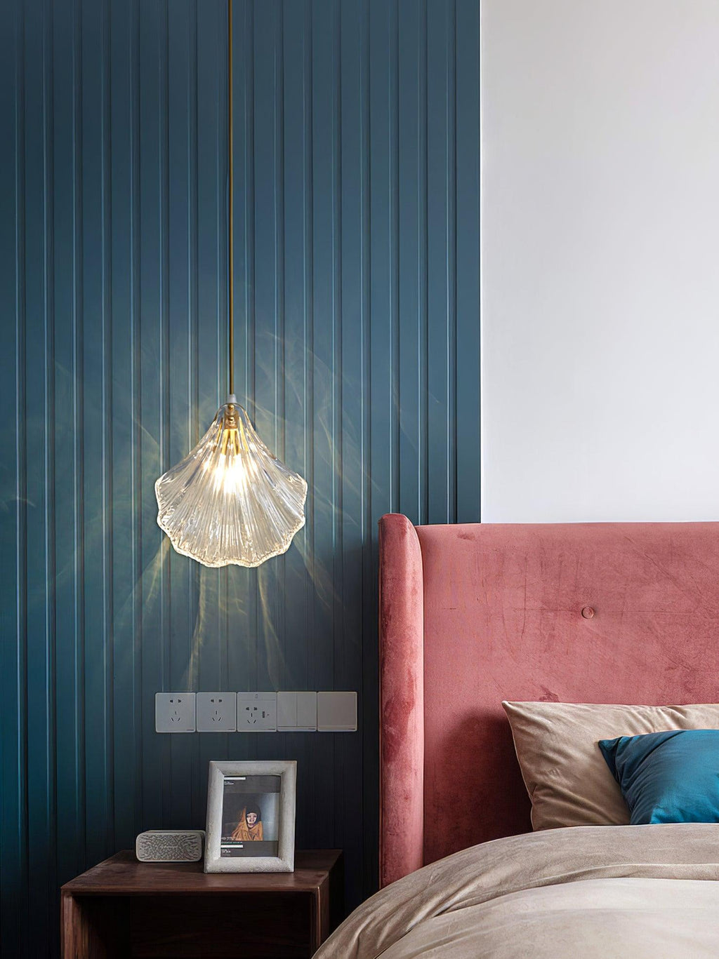 A Clérachic clear glass shell pendant hangs against a teal slatted panel, above a bedside table with a framed photo.