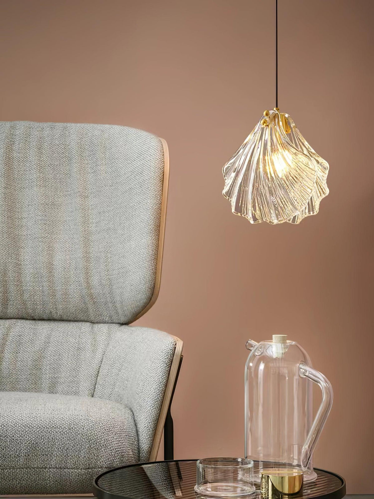 A Clérachic ribbed glass pendant hangs beside a grey armchair and a clear glass jug, against a dusty-pink wall.