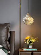 A Clérachic shell pendant in cognac-toned glass lights a grey wall beside a leather headboard and a bunch of yellow flowers.