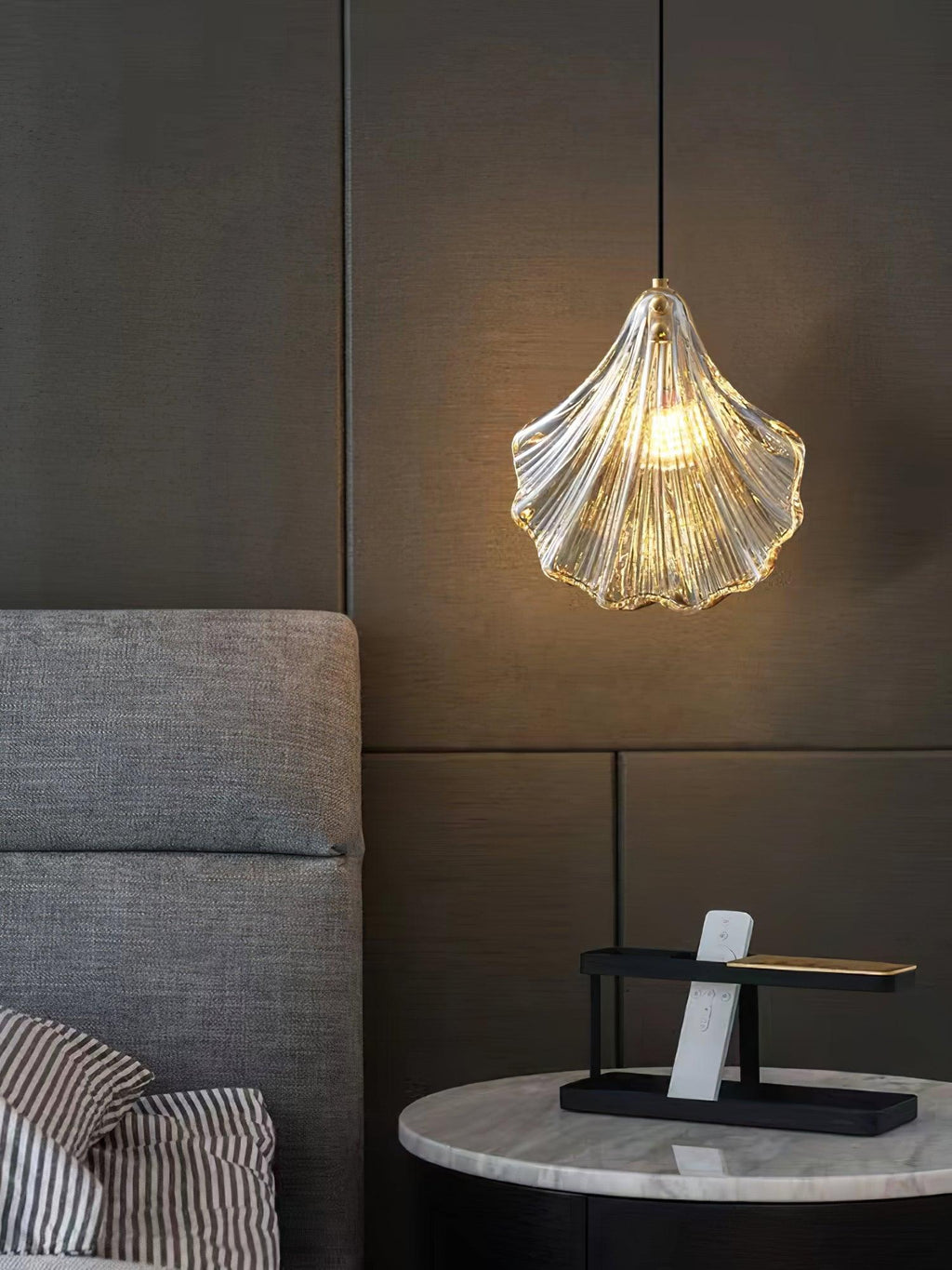 A Clérachic shell pendant glows warm gold above a marble side table, in front of a grey upholstered headboard and dark panelling.