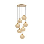 A Clérachic cluster of seven cognac glass shell pendants hangs at staggered lengths from a round brass plate, on white.