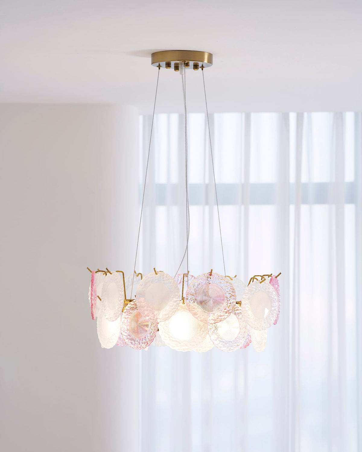 A Clérachic ring pendant glowing in a bright room, with sheer curtains behind it and a table below.