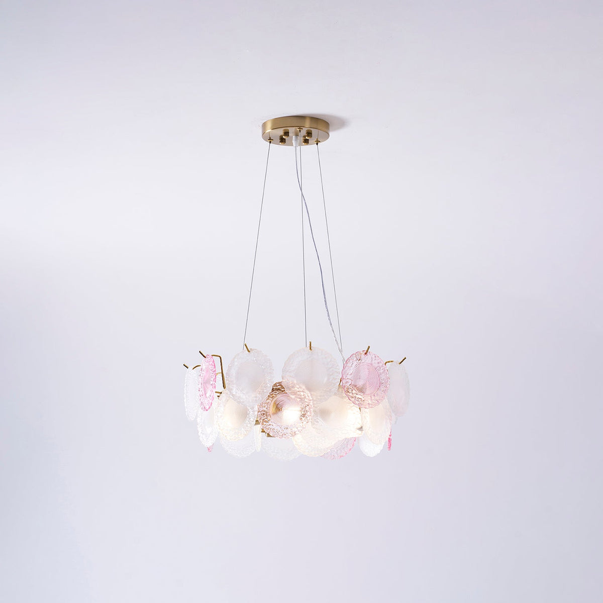 A Clérachic pendant with pink and frosted glass discs, lit, hung on three wires against a pale lilac wall.