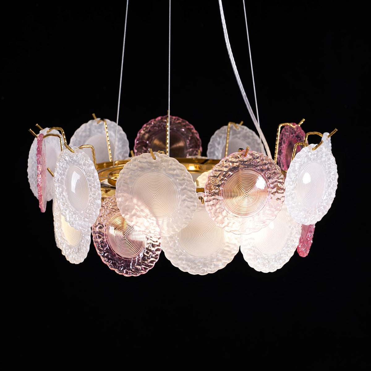 This Clérachic pendant lit on a black backdrop, with pink and frosted glass discs on a gold ring and two white wires above.