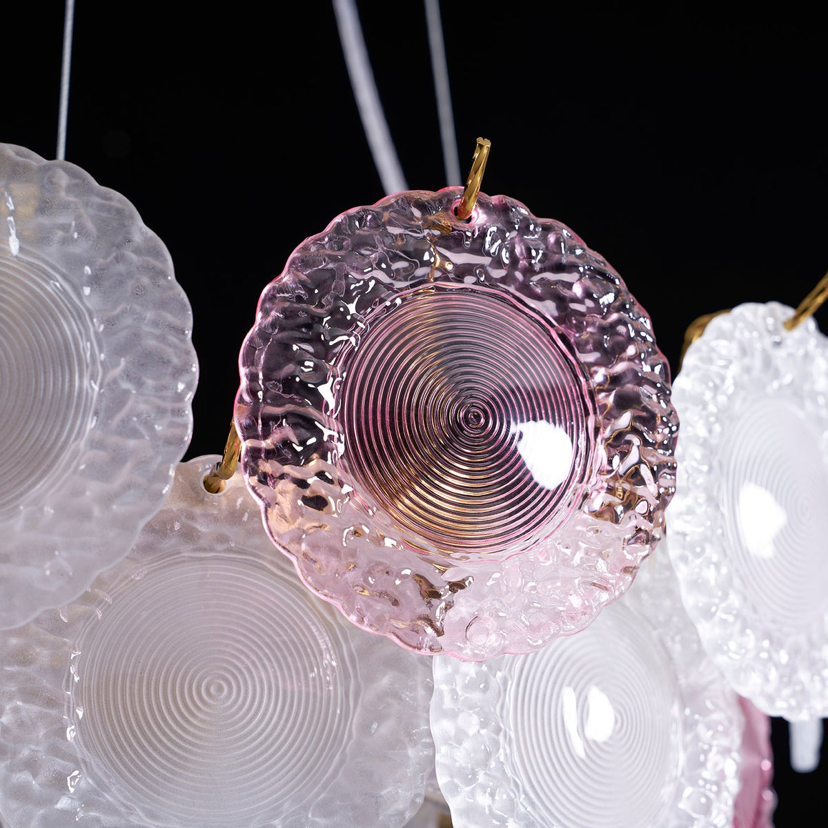 Close view of a pink disc with a ridged centre beside a clear frosted disc, against a dark backdrop, on this Clérachic pendant.