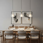 Three Clérachic glass pendants hung in a row above a walnut dining table with grey upholstered chairs, in front of two abstract prints.