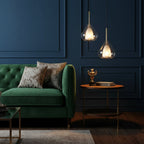 Two Clérachic glass pendants at different heights against a navy panelled wall, above a green velvet sofa and a round copper side table.