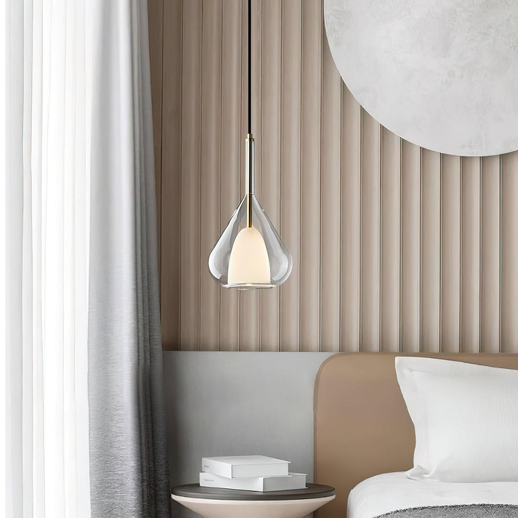 Clérachic clear glass pendant lit in front of a ribbed beige wall panel, beside a bed with white pillows and a stack of books on a side table.