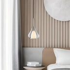 Clérachic clear glass pendant lit in front of a ribbed beige wall panel, beside a bed with white pillows and a stack of books on a side table.