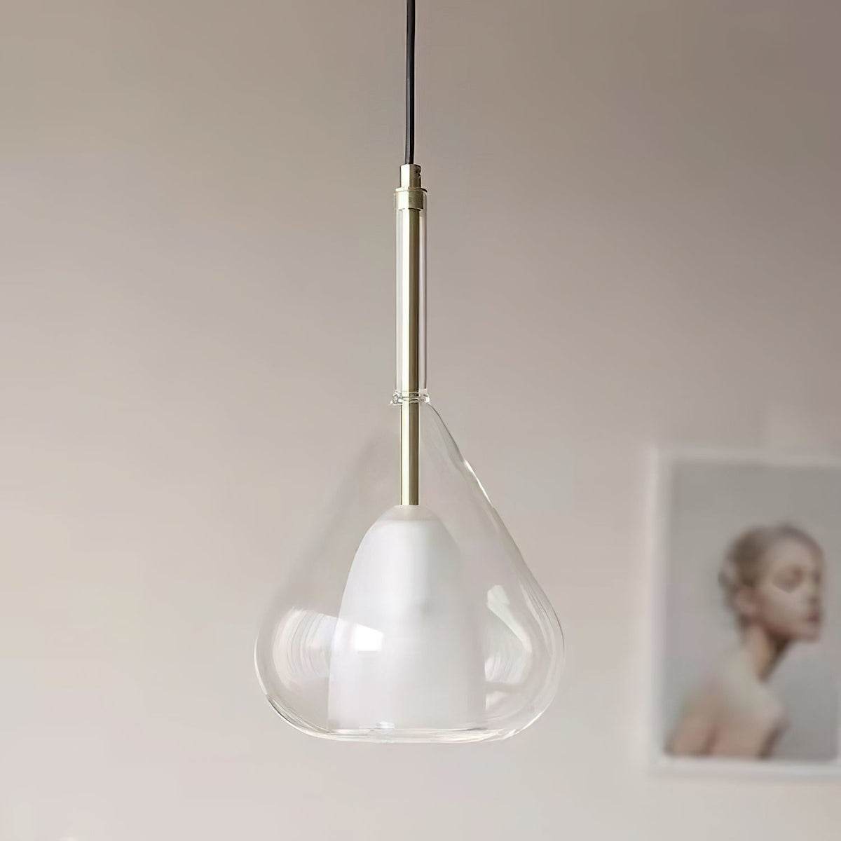 Close view of a clear glass teardrop pendant from Clérachic, with a frosted inner shade, a brass stem and black cord, against a pale wall.