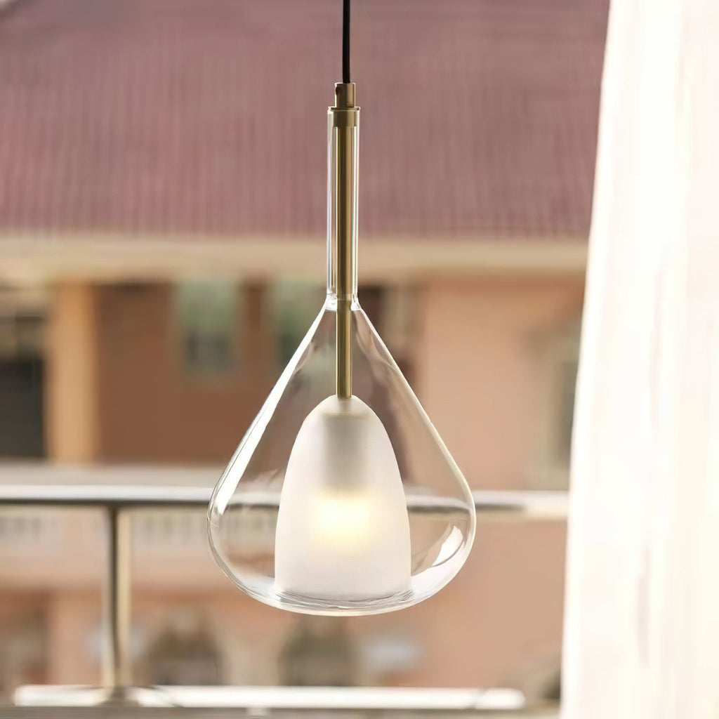 Clérachic clear glass pendant with a brass stem lit by a window, with a blurred terrace and tiled roofs outside.