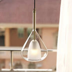 Clérachic clear glass pendant with a brass stem lit by a window, with a blurred terrace and tiled roofs outside.