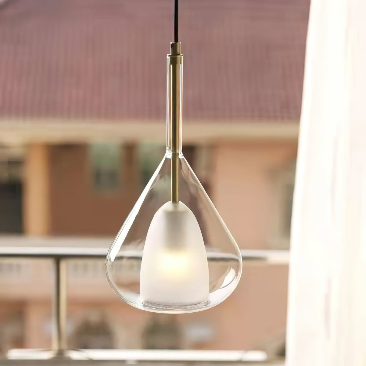 Clérachic clear glass pendant with a brass stem lit by a window, with a blurred terrace and tiled roofs outside.