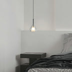Clérachic glass pendant lit above a bedside table with a dark grey bed, hanging against a white wall in a minimal bedroom.