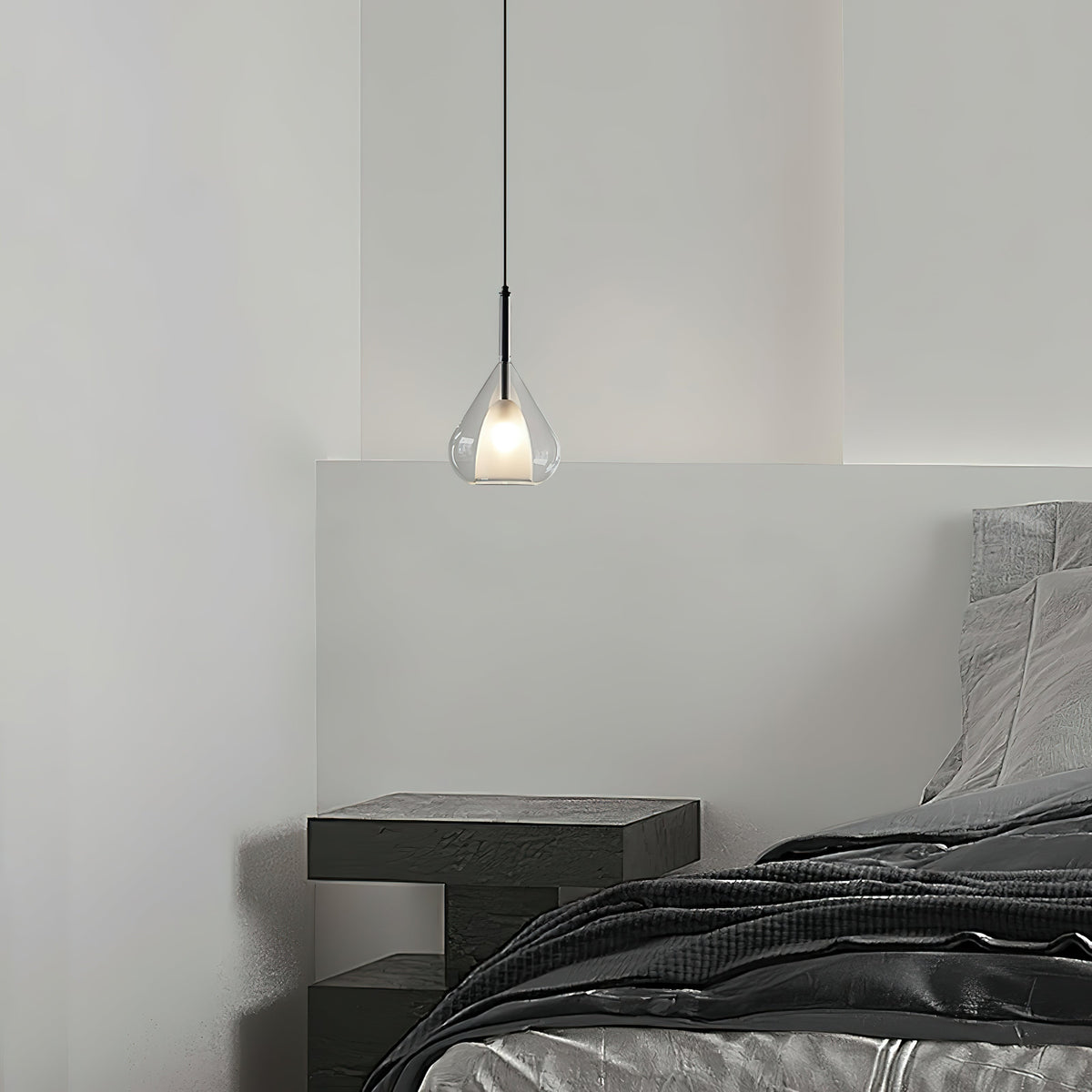 Clérachic glass pendant lit above a bedside table with a dark grey bed, hanging against a white wall in a minimal bedroom.