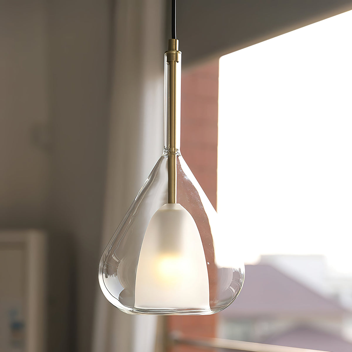 Low angle on a Clérachic clear glass pendant beside a bright window, its frosted inner shade glowing softly above a brass stem.