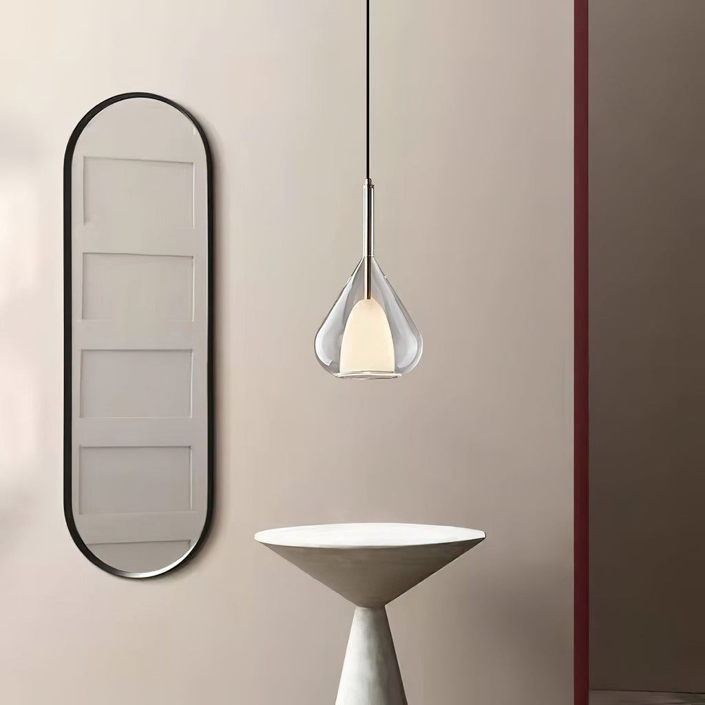 Clérachic clear glass pendant lit beside a tall black-framed oval mirror and a pale conical side table, on a blush wall.
