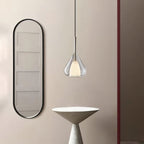 Clérachic clear glass pendant lit beside a tall black-framed oval mirror and a pale conical side table, on a blush wall.