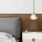 Clérachic clear glass pendant hanging above a bedside table with a white drawer and brass pull, next to a grey headboard on a bronze panel.