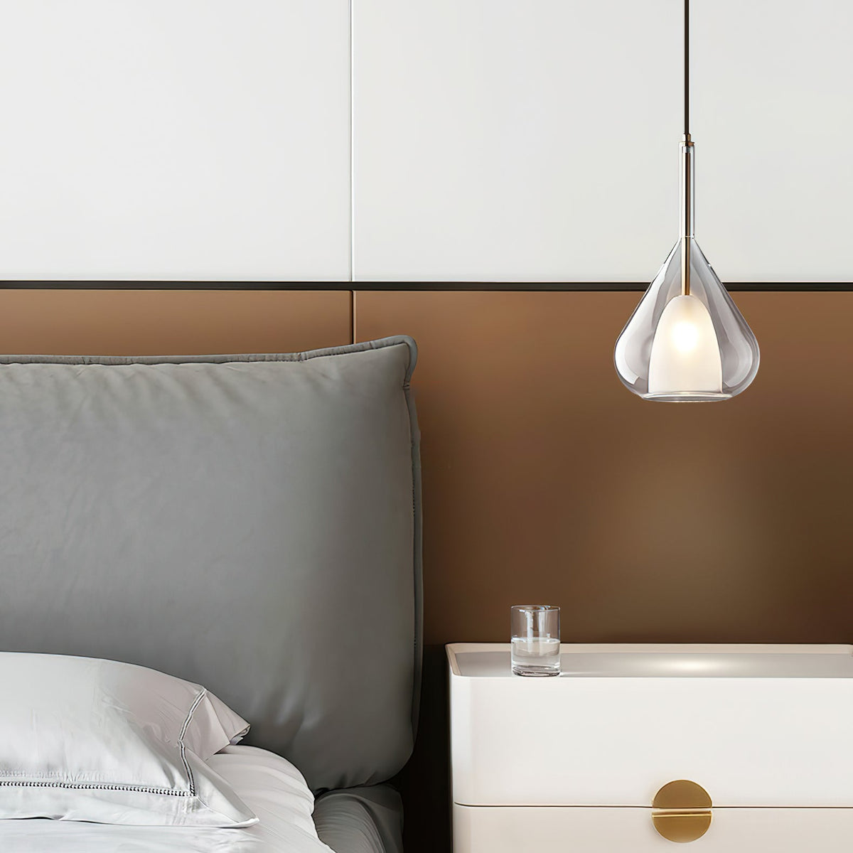 Clérachic clear glass pendant hanging above a bedside table with a white drawer and brass pull, next to a grey headboard on a bronze panel.