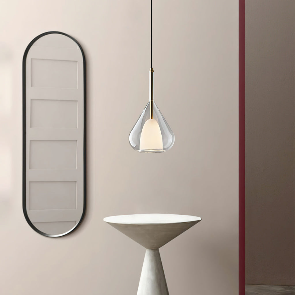 Clérachic clear glass pendant lit beside a tall black-framed oval mirror, with a pale conical table below, seen from a slightly wider angle.