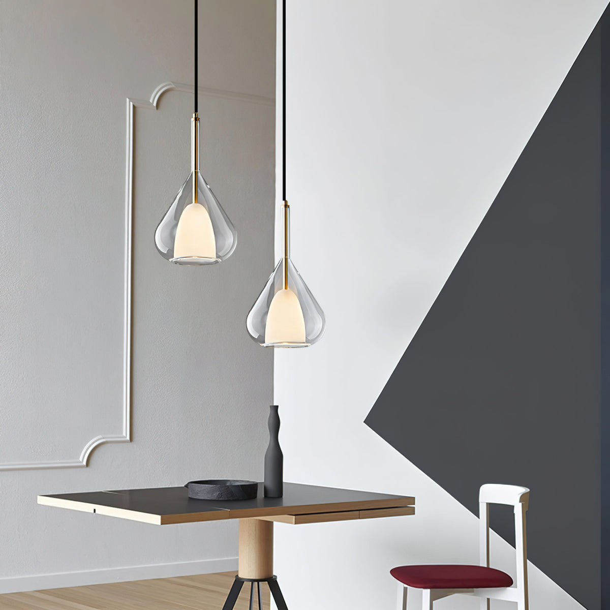 Two Clérachic clear glass pendants at different heights in a room with grey wall mouldings and a dark angular panel, above a small table.
