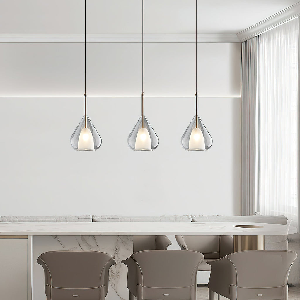 Three Clérachic clear glass pendants in a row above a marble-topped counter with taupe chairs, beside sheer curtains.