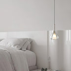 Clérachic clear glass pendant lit above the foot of a pale bed, with soft white bedding against a white wall.