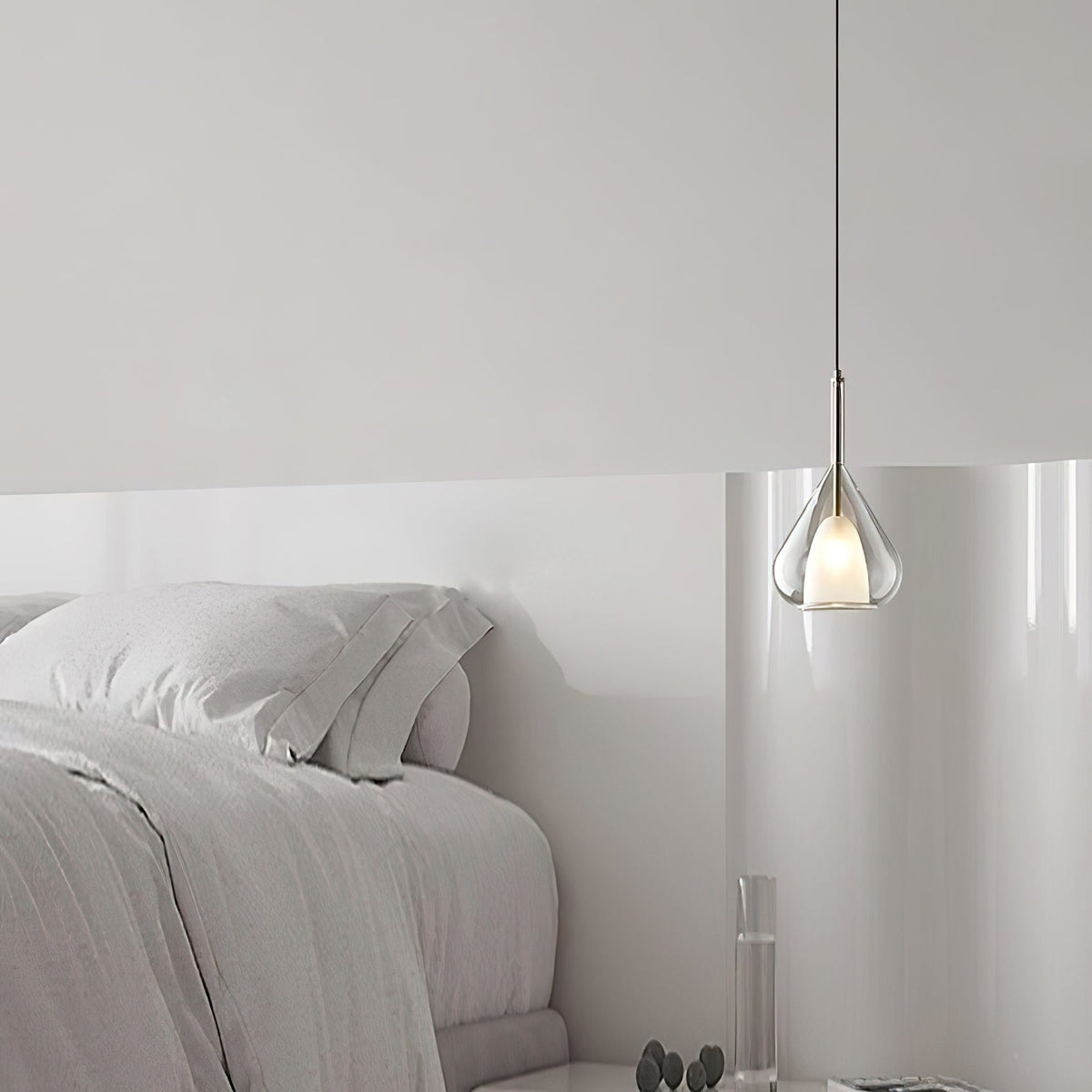 Clérachic clear glass pendant lit above the foot of a pale bed, with soft white bedding against a white wall.
