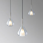 Three Clérachic clear glass pendants at staggered heights against a graduated grey backdrop, each lit with a warm frosted inner shade.
