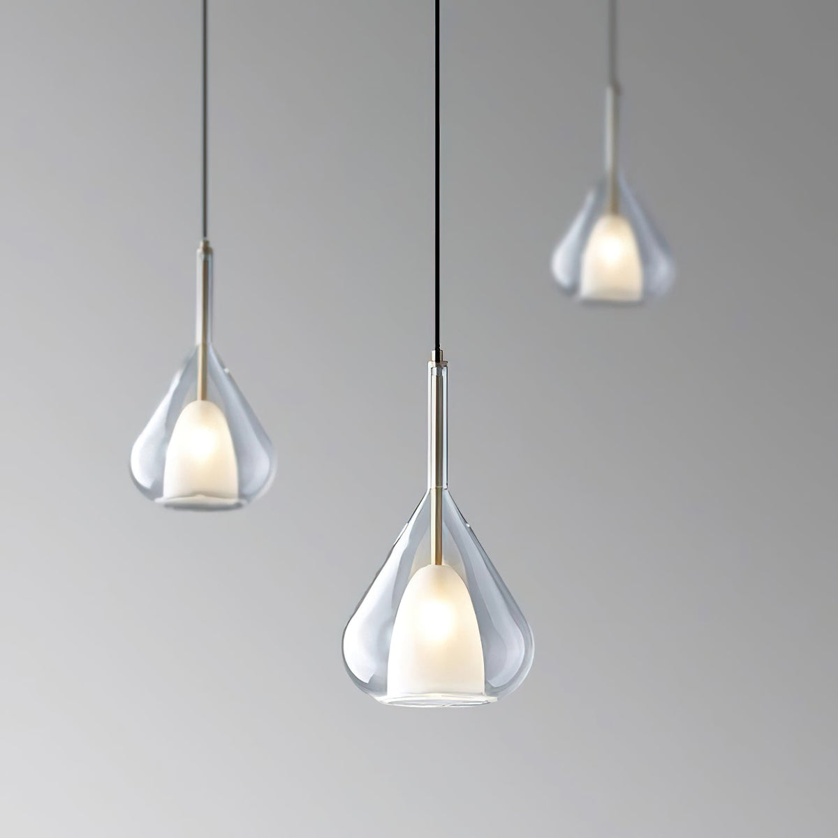 Three Clérachic clear glass pendants at staggered heights against a graduated grey backdrop, each lit with a warm frosted inner shade.
