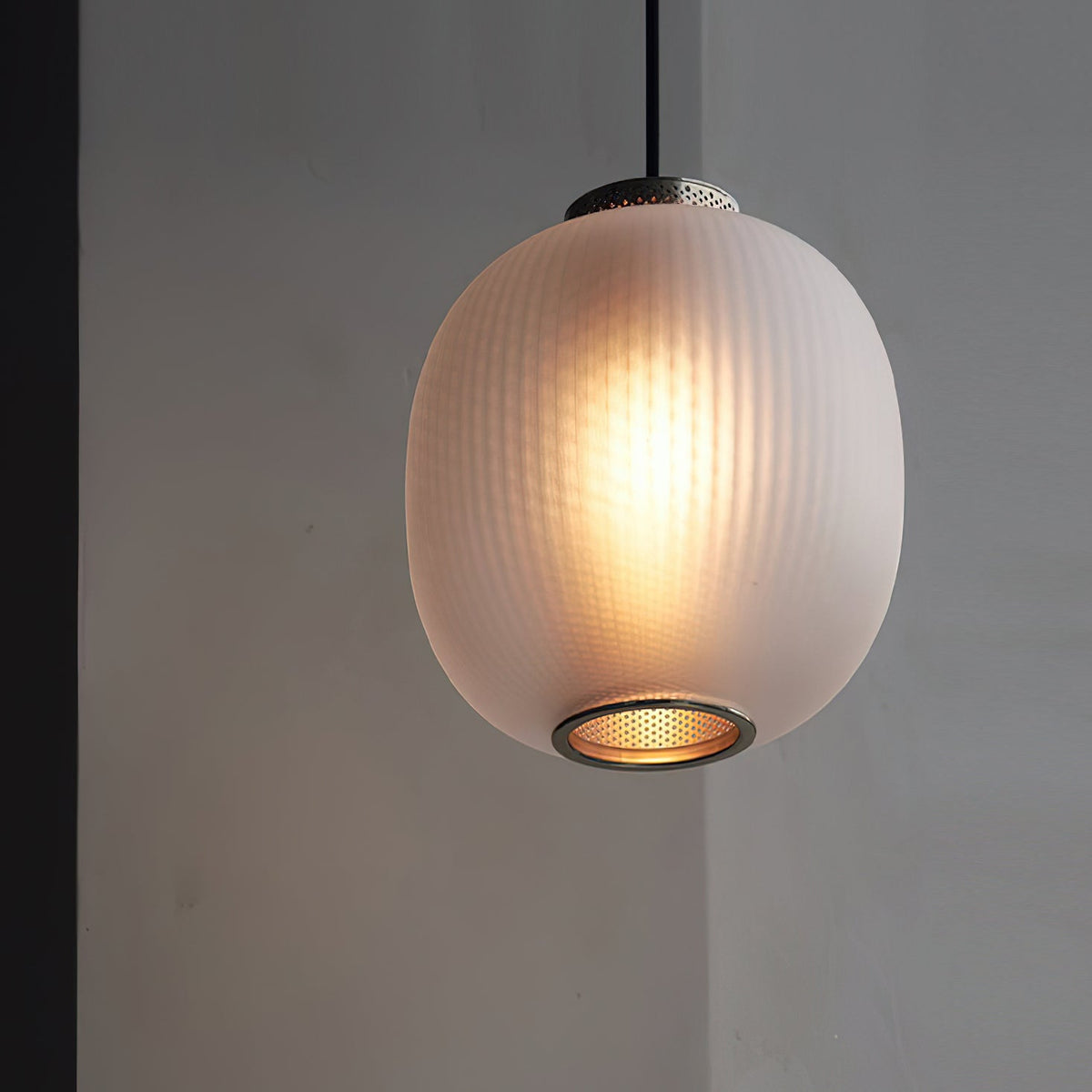 Close view of a lit matte white ribbed glass shade on this Clérachic pendant, with a perforated metal ring at the base, against a grey wall.