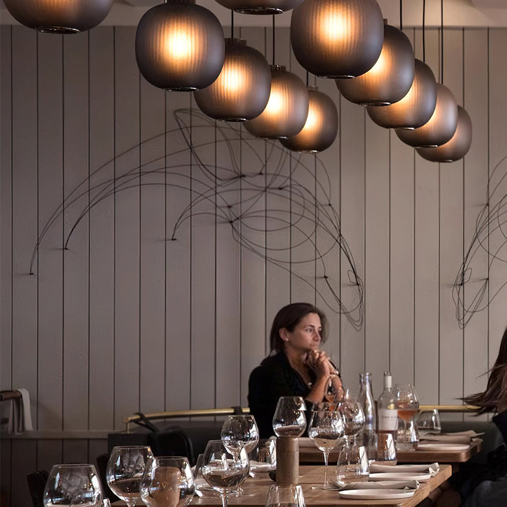 Rows of smoke-grey Clérachic glass pendants glowing over a restaurant, with a diner at a long wooden table below.