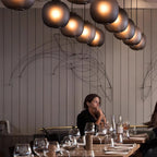 Rows of smoke-grey Clérachic glass pendants glowing over a restaurant, with a diner at a long wooden table below.