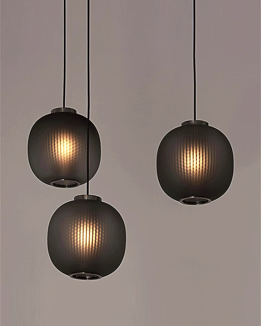 A trio of smoke-grey Clérachic pendants at staggered heights against a taupe wall, light showing through the fine vertical ribbing.