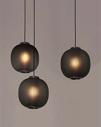 A trio of smoke-grey Clérachic pendants at staggered heights against a taupe wall, light showing through the fine vertical ribbing.