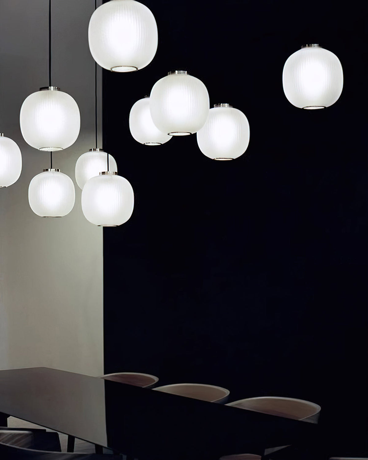 Cluster of matte white Clérachic glass pendants glowing against a dark wall, above a long dark table with chairs.
