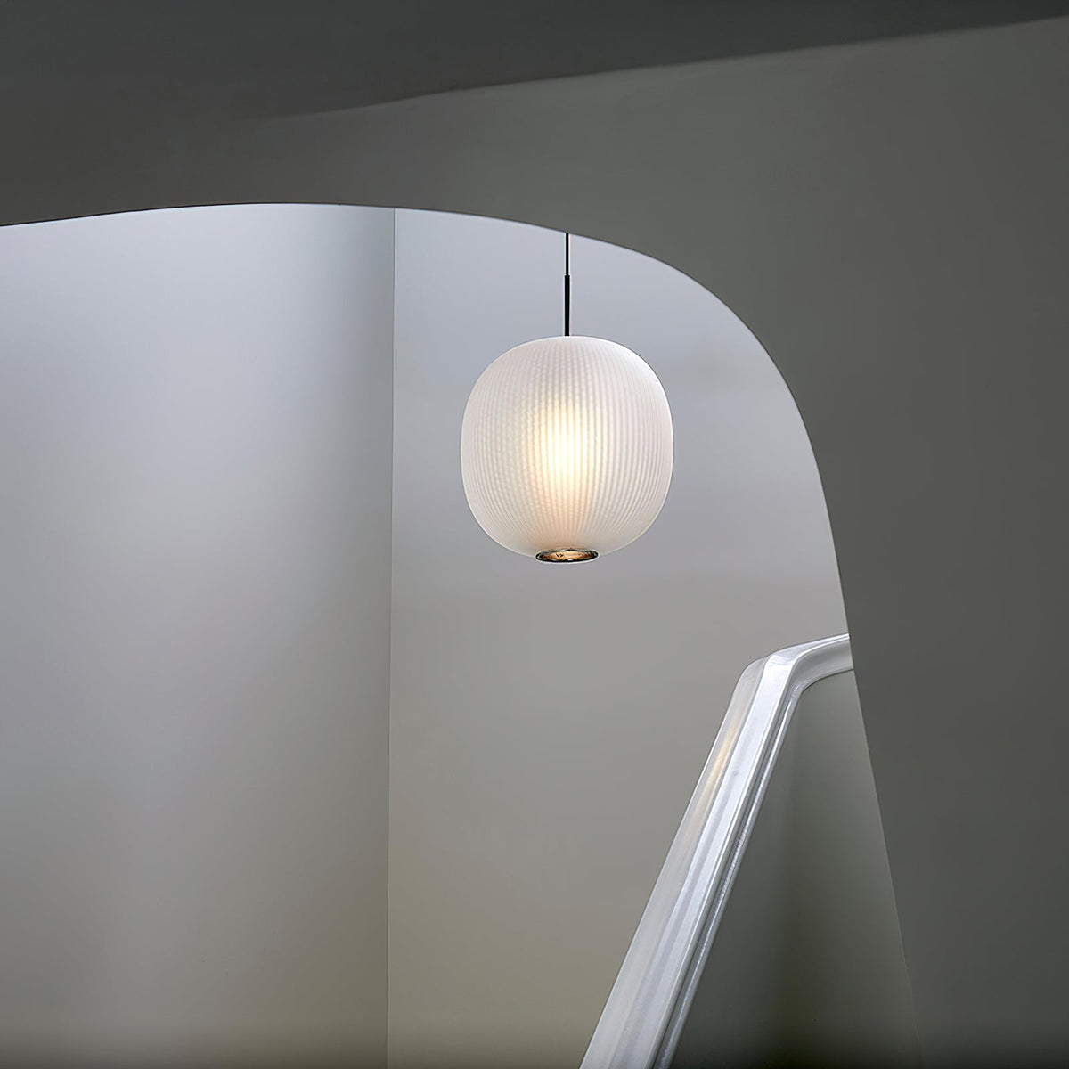Clérachic matte white ribbed pendant hanging in a pale stairwell beside a curved white handrail.