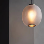 Close view of a lit matte white ribbed glass shade on this Clérachic pendant, warm light glowing inside and a metal mesh ring below.