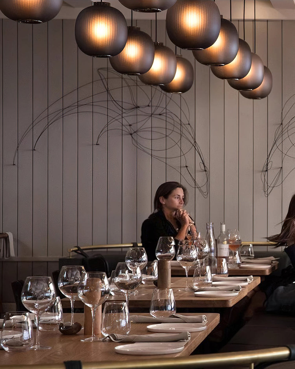 Smoke-grey Clérachic glass pendants in a long curving row above a restaurant, with a seated diner at a wooden table.